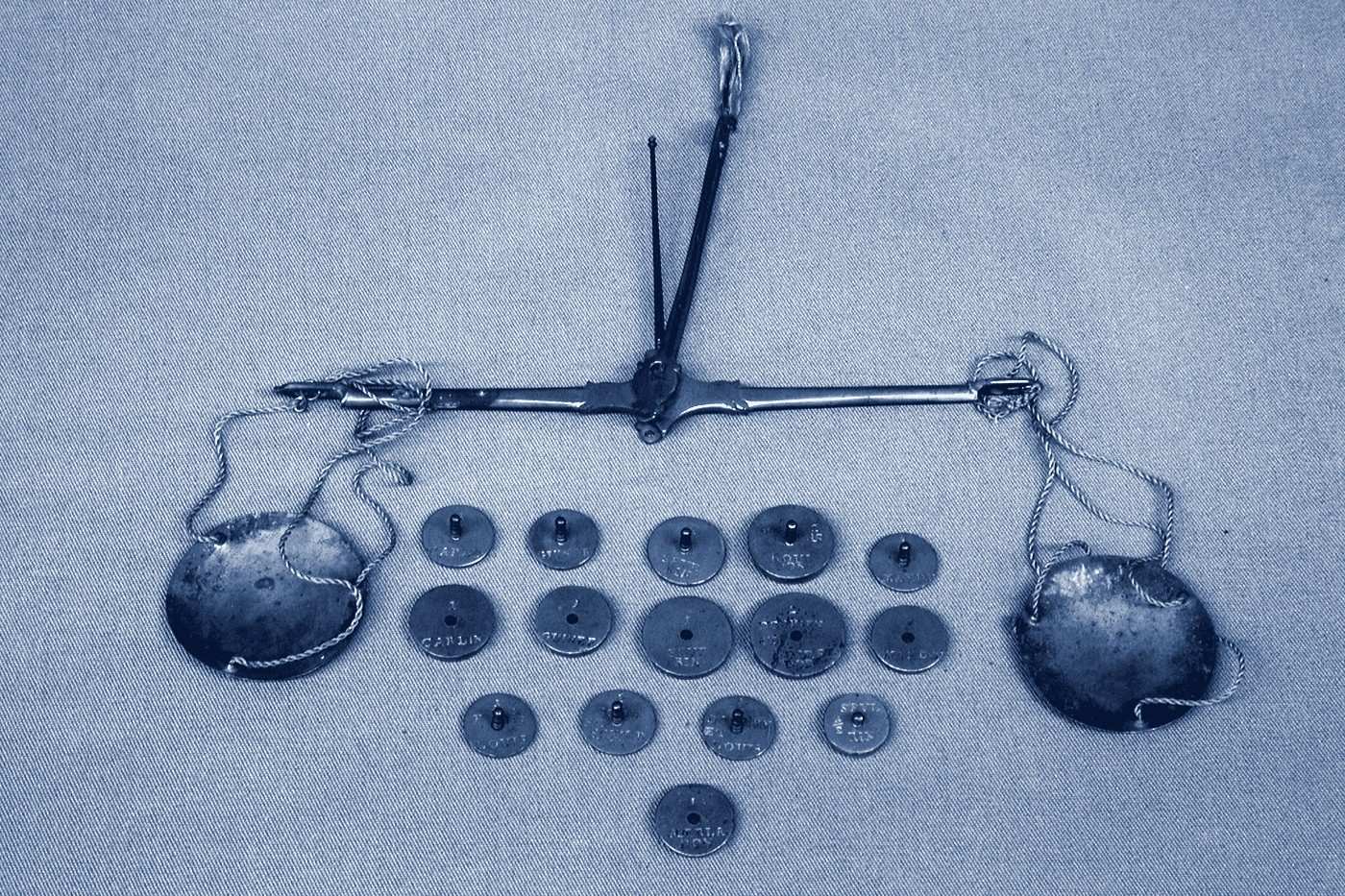 A small brass hand balance laid flat with its two pans and a set of round coin weights.